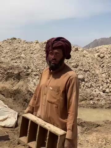 The video shows a man in a brown tunic (qamis) and a maroon turban, with a dark beard, standing outdoors. He holds a multi-compartment mold, likely used for making bricks or blocks, and appears to be speaking or explaining something. To his left, another person is partially visible, kneeling or squatting near a white tarp on the ground.

The setting is an open, arid area with large mounds of earth, dirt, and rock fragments in the background, suggesting an excavation or processing site. A small pool of muddy water is visible on the ground to the right. Distant hills or mountains rise under a bright, clear sky.

The activity taking place appears to be related to the manual production of construction materials, specifically bricks or blocks, given the presence of the mold, raw earth, and water. The bright sunlight indicates it is daytime, with clear weather conditions. The attire and landscape are consistent with the provided location context of Deh-E Bagh, Afghanistan. There is no visible text.