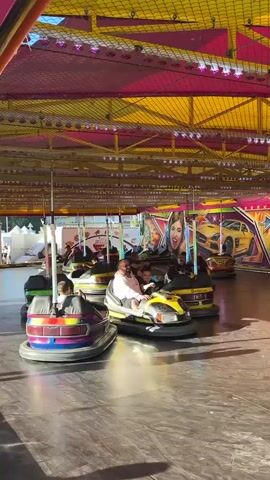 A bumper car ride at a carnival is depicted. The ride has a brightly colored canopy overhead, with yellow and pink fabric stretched across a metal frame, adorned with strings of lights. The bumper cars themselves are varied in color and design, with some being yellow, blue, and red. Several people are seated inside the cars, some driving and others as passengers. The cars are in motion, bumping into each other on a flat, shiny surface. In the background, there are large decorative panels featuring images of women, sports cars, and stars, adding to the festive atmosphere of the amusement park. The lighting appears to be a mix of natural light and the artificial lights strung across the canopy.
