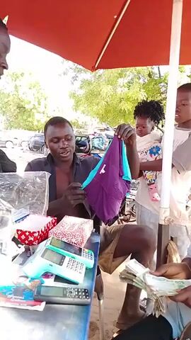 A man is selling underwear at an outdoor market. He holds up a pair of purple and blue underwear to show a customer. The vendor is sitting behind a blue table with several items including two blue card readers, money, and various merchandise. Two other people stand by the table looking at the merchandise. The scene is sunny and outdoors under a red umbrella.