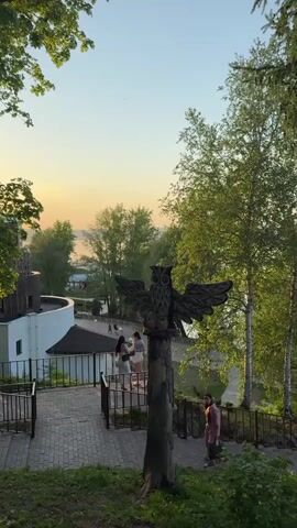 The scene is an outdoor public space with a landscaped park area on a slope leading down to a body of water, likely a lake or a wide river. The time appears to be late afternoon or early evening, indicated by the soft lighting and hint of sunset colors on the horizon.  

In the foreground, there is a decorative wooden sculpture of an owl with outstretched wings, mounted on a tree stump. A paved walkway, constructed with brick, descends the slope. Several people are visible walking along the walkway and near the bottom of the stairs.

A woman in a pink dress is walking down the stairs, while two women, one in a white crop top and the other in a dark top, walk towards the viewer, past the owl sculpture.

The environment is green, with trees framing the scene. A modern, white-walled building with a dark roof is visible at the top left. The body of water in the background stretches across the horizon, adding depth to the scene.  The sky is clear with a soft gradient of color towards the horizon. Lamp posts are placed along the lower walkway near the water.