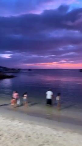 The video shows a beach scene at dusk. The sky is filled with vibrant colors of purple, pink, and orange, with dark clouds scattered across it. The ocean reflects the colors of the sky, appearing in shades of deep purple and pink. Several boats are visible in the distance on the water. In the foreground, on the sandy beach, two people are partially visible. One person appears to be an adult wearing a white shirt, and the other is a child. They are standing in shallow water near the shore. The camera is positioned on the beach, looking out towards the ocean and the sunset.