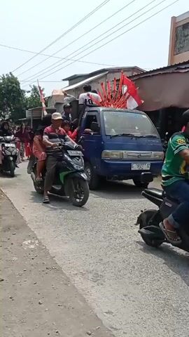 A group of people are participating in a parade or procession. Several individuals are riding on the back of a blue truck, some holding red and white flags. A man and a woman are riding on a motorcycle in the foreground, with the man driving and the woman sitting behind him. Another motorcycle with two people is visible to the left, and more people, including children, are walking and riding on motorcycles in the background. The procession is taking place on a paved road, with buildings and trees visible on either side. The overall atmosphere appears festive and celebratory.
