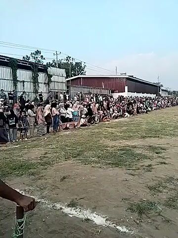 A soccer game is being played on a dirt field with patchy grass. A large crowd of people is gathered along the sideline, some sitting and some standing, watching the game. The crowd is concentrated on the left side of the field, forming a long line. There are buildings and trees in the background. The sky is mostly cloudy. The video pans across the scene, showing the crowd and the game in progress.