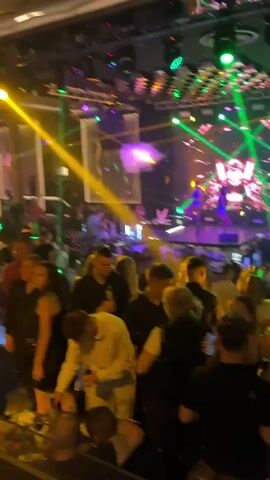 A crowded nightclub is visible in this video. The room is dark, illuminated by colorful stage lights and a large LED screen at the back displaying abstract patterns and imagery. People are gathered on the dance floor and at tables, with many dancing or conversing. A DJ booth is situated on a raised platform behind the crowd, with two DJs visible. Smoke or fog machines are active, adding to the atmosphere. The overall impression is one of a lively party or club event.