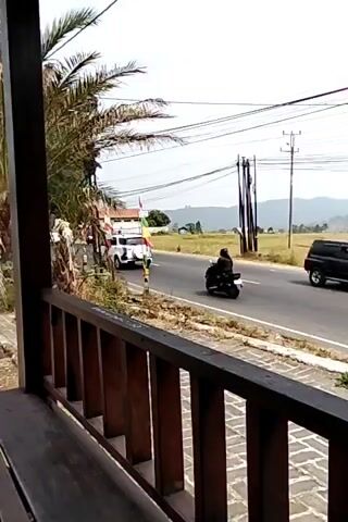 The video begins with a view from behind a wooden railing, looking out at a road with several motorcycles and a truck. Palm trees are visible on the left side of the screen, along with a building and some flags. The road is asphalt, with a yellow line down the center. Fields of dry grass are on the right side of the road, with power lines and poles in the background. The sky is overcast. Several motorcycles are seen traveling down the road. One motorcycle in the distance has a very tall structure attached to it. A large, yellow flatbed truck is also seen driving down the road. The video captures a moment in time on a rural road, with traffic and a natural landscape.