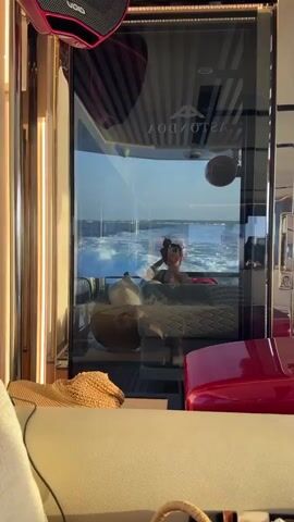 The video shows a reflection in a boat window. A person is visible in the reflection, looking at their phone while on the boat. Outside the window, the ocean is visible with waves. Inside the boat, there are pillows and other furniture. The lighting suggests it is either sunrise or sunset.
