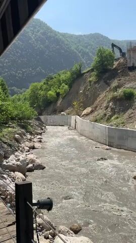 A wide, murky river flows through a mountainous landscape. Concrete walls line the right bank of the river, forming a channel. On the steep, wooded slope above the right bank, construction is underway. An excavator is visible, along with unfinished building structures. The left bank of the river is rocky and less developed, with trees and foliage growing down to the water's edge. The sky is clear and blue, indicating a sunny day. The camera is positioned low, possibly from a bridge or deck overlooking the river.