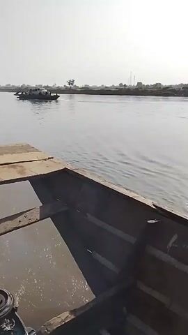 A boat moves along a river, visible in the background is another boat moving in the same direction. The camera angle is from the inside of the boat, looking out, then pivots down to reveal the engine and the legs of a person steering the boat. A yellow fuel tank is on the floor of the boat.