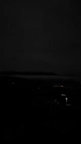 The video shows a dark, nighttime scene with a distant cityscape visible in the horizon. There are faint lights from buildings or vehicles, and the sky is overcast with clouds. The foreground appears to be a dark, elevated area. A leafless tree is visible on the right edge of the frame.