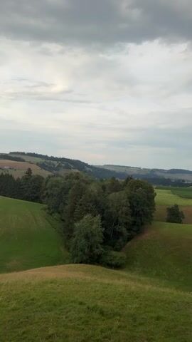 This is a vertical video with a landscape panorama showing rolling green hills, trees, and a small village. The sky is overcast with dramatic, dark clouds gathering, suggesting an impending storm. The video begins with a view of the hilly countryside with fields and forests. As the video progresses, the camera pans to reveal a village nestled on a hilltop, with houses and a church steeple visible. The latter part of the video focuses on a section of a building's exterior, including a roof, gutter, and a balcony or patio area with railings, giving a sense of proximity to a residential or hospitality setting. The overall mood is serene yet somewhat foreboding due to the weather. The time of day appears to be late afternoon or early evening. There is no visible text in the media. The location is Ufhusen, Switzerland.