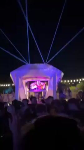 Here's a concise description of the video:

The video shows an outdoor music event at night. A crowd of people is gathered in front of a stage that is illuminated with bright blue and purple lights. Above the stage are long blue laser beams extending upward. A DJ appears to be performing on stage, and the crowd is facing them and dancing. An inflatable green alien toy floats above the crowd. String lights are visible in the background.