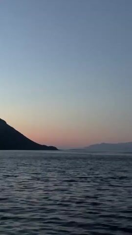 The video shows a body of water with gentle waves, likely a sea or a large lake, at dusk. The sky transitions from a deep blue at the top to a lighter pastel hue near the horizon, with hints of pink and orange, indicating the sun has recently set or is about to rise. To the left, a silhouetted mountain range dominates the landscape, its dark mass contrasting with the lighter sky. Further in the distance, another mountain range is visible, appearing fainter and more subdued. The overall atmosphere is serene and tranquil, with no signs of human presence or activity.