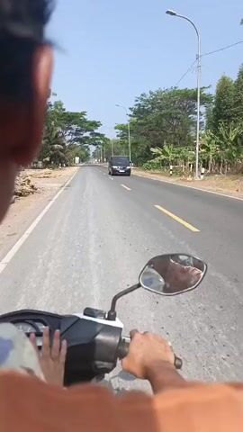 The video shows a first-person perspective of someone riding a motorcycle on a paved road under a clear sky. In the foreground, the rider's hands are visible on the handlebars. The motorcycle has a black mirror reflecting a person's face. The road is bordered by green trees and vegetation. Further ahead, other vehicles, including a black van and two motorcycles, are visible traveling in the same direction. The road has yellow lane markings.