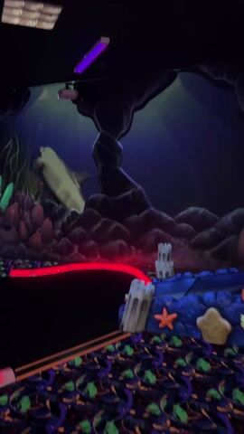 The video shows a glow-in-the-dark miniature golf course. The foreground is a black carpet with neon purple and green swirling designs. The miniature golf hole in the foreground has two stone-look castle decorations, an orange starfish, and a beige star. A red light spans across the center of the scene. The background consists of a rock formation in the center, with painted coral and plant life to the left, and an animated shark projected on the wall. A projection of an octopus is added to the right-hand side of the scene after 3 seconds.