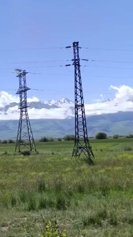 A stop sign is visible in the foreground, partially obscuring the view of a vast grassy field. In the middle ground, two tall electrical towers stand prominently, connected by wires that stretch across the frame. Beyond the towers and the field, a range of mountains is visible, their peaks partially obscured by clouds. The sky is a clear blue, indicating a sunny day. The overall impression is one of a rural landscape with evidence of human infrastructure.