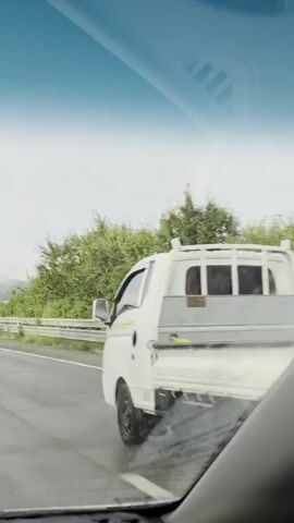 The video shows a view from inside a car driving on a wet road, likely a highway, under a cloudy sky. A bright rainbow arcs across the sky in the distance, partially obscured by trees and a hill. The car's windshield is visible in the foreground, with a reflection on its surface. A concrete barrier and a metal guardrail line the side of the road. Lush green trees and foliage are visible beyond the guardrail, and distant mountains can be seen through the haze. The road markings, including white lines, are clear. The scene suggests a recent rain shower, given the wet road surface. There are no people or other vehicles clearly visible in the frame.