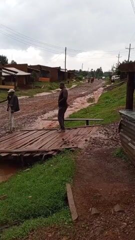 The video shows a muddy dirt road in what appears to be a rural village. Several buildings with metal roofs are visible on either side of the road. The ground is a mix of red dirt and green grass, and puddles of muddy water cover much of the road. Several people are walking on the road. At the start of the video, three men are standing on a wooden bridge or platform spanning a small waterway. The sky is overcast and gray. In the background, a light pole rises above the trees. The camera initially focuses on the men on the bridge before panning across the landscape, revealing more of the village scene.