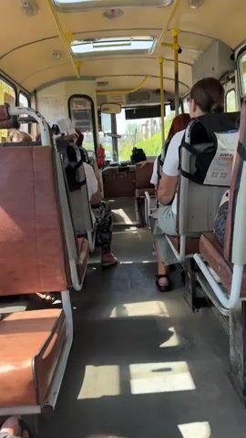 The video shows the interior of a bus, likely an older model, with several passengers seated. The seats appear to be upholstered in a brown vinyl material and are arranged in rows facing forward. Some of the seats are empty, while others are occupied by passengers. The floor of the bus is a dark color, possibly linoleum or rubber. The ceiling is light colored with several overhead compartments and vents. Yellow handrails are visible running along the ceiling and on the back of some seats. Sunlight streams in through the windows, casting shadows on the floor. The overall impression is of a functional but somewhat aged public transportation vehicle.