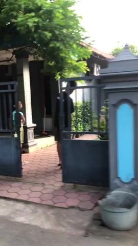 The video shows an exterior view of a house. Two people are visible, one standing near the gate and another inside the yard. The gate is partially open. A tree is growing near the house. A broom is leaning against the wall. The ground is paved with hexagonal tiles.
