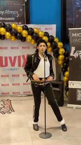 This is a video of a Michael Jackson impersonator performing on a stage. The performer is a person dressed in a black jacket, white shirt, and black pants, mimicking Michael Jackson's iconic style. They are holding a microphone and appear to be singing and dancing.

The backdrop features a balloon arch in black and gold, and a banner with the text "MICHAEL JACKSON" and an image of the singer. There are also logos for "MUVIX CINEMAS". The floor is tiled, and a speaker is visible on the right. In the background, a movie poster is displayed. A young child can be seen near the stage on the right side of the frame.

The setting appears to be an indoor event, possibly at a cinema or a public gathering place, given the "MUVIX CINEMAS" branding. The atmosphere seems festive and energetic, indicated by the performance and the balloon decorations. The lighting suggests it could be daytime or a well-lit indoor venue. The overall impression is that of a tribute performance or a promotional event related to Michael Jackson.