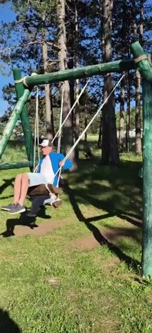 Two individuals are seen on a wooden swing set. The swing set is painted green and has a sturdy wooden frame. The ground beneath the swing set is covered in green grass. In the background, tall trees with thin trunks are visible, suggesting a wooded area. Sunlight filters through the trees, casting shadows on the ground.

The individual on the left, wearing a dark jacket and grey pants, is seated on a wooden swing suspended by ropes. They are wearing a white cap.

The individual on the right, wearing a blue and white t-shirt and light-colored shorts, is also seated on a wooden swing suspended by ropes. They are wearing a white cap and dark sneakers.

Both individuals appear to be swinging. The person on the right is swinging higher than the person on the left. The scene is captured in daylight under a clear sky.