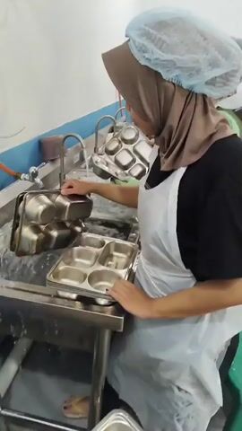 A person wearing a hijab, hairnet, and apron is washing metal trays in a sink filled with soapy water. There are several stacked metal trays on the counter in front of the person. Another person is visible in the background, also washing trays. The environment appears to be a commercial kitchen or food service area with stainless steel sinks and counters.