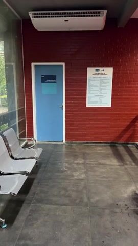The video pans across a waiting area with a red brick wall. To the left, a blue door is marked with "Epidemiologia" and an arrow pointing left. A white sign with text and a logo hangs on the brick wall to the right of the door. In the foreground, rows of empty, metallic chairs are visible. An air conditioning unit is mounted on the wall above the door. The floor is tiled in a dark gray. The video ends with a shot of a railing on the right side of the frame.