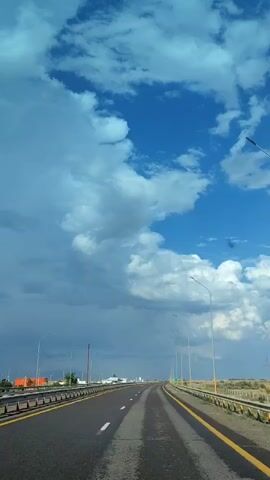 The video shows a view from inside a vehicle traveling on a highway. The sky is filled with dramatic, billowing clouds, transitioning from bright blue to dark gray, suggesting an impending storm or the aftermath of one. The road ahead is empty and appears wet in patches, indicating recent rain. Yellow lines mark the edges of the lanes and the shoulder. Along the sides of the highway, there are streetlights and low vegetation. In the distance, some buildings and structures are visible, as well as a few power poles. The overall impression is one of a vast, open landscape under a dynamic sky.