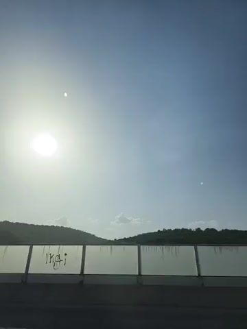 The video shows a bright, sunny sky with the sun visible as a large, bright circle in the upper left quadrant. Two small, faint dots are also visible in the sky. Below the sky, there is a line of hills covered in green foliage, with a translucent barrier running horizontally across the lower portion of the frame. The barrier appears to be made of glass or a similar material with vertical supports. The very bottom of the frame shows a dark surface, possibly a road or ground. The scene suggests an outdoor environment, likely during the daytime, with a highway or elevated viewpoint given the presence of the barrier. There are no people or distinct objects other than the natural landscape and the barrier. The lighting is bright, indicating a sunny day.