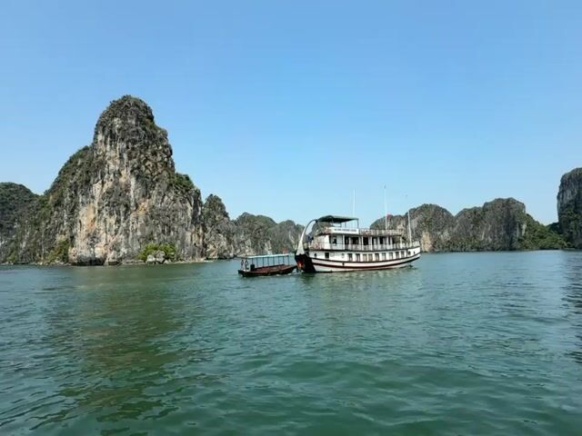 The video presents a serene daytime scene in a bay, characteristic of Ha Long Bay in Tuan Chau Ward, Vietnam. The main subjects are two boats and numerous towering limestone karsts rising from the water under a clear blue sky.

In the foreground, an elegant, multi-deck cruise boat, predominantly white with red accents, is visible on the calm, emerald-green water. Partial text on its side reads "HALONG" and "PREMIUM CRUISE." The boat features an upper deck with an awning and multiple windows along its lower decks. A smaller, dark-colored boat, likely a tender or shuttle, is docked closely alongside the larger vessel, suggesting a passenger transfer point or preparation for an excursion.

The backdrop is dominated by majestic limestone karsts, typical of the region, covered in vibrant green foliage. A large, distinct karst stands prominently on the left side of the frame, showcasing its rugged, textured rock face. Other karsts are scattered towards the horizon, creating a dramatic and picturesque landscape.

The water is tranquil, with gentle ripples reflecting the clear, bright blue sky, which is devoid of clouds. The bright sunlight indicates a pleasant day. The gentle hum of a boat motor can be heard throughout the video, contributing to the peaceful atmosphere of the open water. The scene evokes a sense of natural beauty and tranquility, indicative of a popular scenic tourist destination.