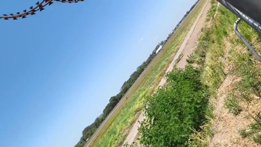The video captures a wide-angle view of an airport runway on a clear, sunny day. The camera is tilted, giving the impression of a dramatic perspective. In the distance, a white airplane with blue accents is visible on the runway, appearing to be either taking off or landing. The runway is bordered by a strip of green grass and a line of trees. In the foreground, to the right, there is dense green vegetation, and to the left, a section of barbed wire fence is visible, suggesting the perimeter of the airport. The sky is a vibrant, cloudless blue. The scene is still and peaceful, with no people directly visible.