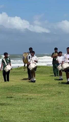 The video shows a group of young males, likely students, dressed in white shirts and dark shorts or pants, practicing with drums and a saxophone on a grassy area near a beach. The ocean with waves is visible in the background, under a partly cloudy sky. A white stupa-like building is situated on a hill to the right of the frame. The individuals are positioned in a loose formation, with some holding and playing drums of various sizes, and one person playing a saxophone. Their actions suggest a rehearsal for a performance or parade. The lighting indicates it is daytime, likely mid-morning or afternoon.