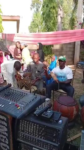 The scene appears to be at an outdoor event, possibly a celebration, featuring live music. Several people are present, including musicians and onlookers. A group of musicians are playing traditional instruments, including drums and a stringed instrument, as others watch and listen. The musicians are seated on chairs, and the musical equipment is set up nearby. A sound mixer, a microphone, and an amplifier are visible in front of the musicians.

The setting is outdoors, with a wall, trees, and a pink cloth draped overhead providing shade. The people are wearing a mix of traditional and casual clothing. There is a portable generator in the background. One man is wearing a green baseball cap, another is wearing a white traditional hat. The camera moves somewhat, showing different angles of the scene.