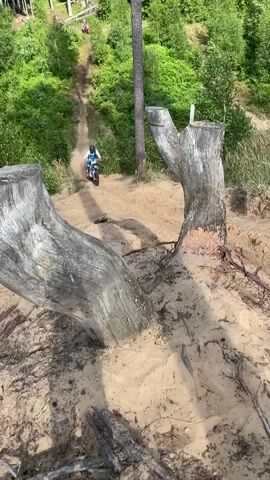 A person rides a dirt bike down a sandy track in a wooded area. The rider is wearing a blue and white dirt bike suit and a black helmet. The dirt bike is silver and black. The track is surrounded by trees and sand. The camera is positioned close to the ground, looking up at the rider.