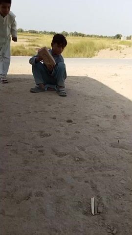 The video opens with a young boy crouching on dusty ground, holding a brick. He is looking down at something in his hand. Behind him, another young boy stands, leaning forward and looking in the same direction. A man rides a motorcycle in the background. The environment appears to be an open, outdoor area with a dirt road and dry vegetation. The video then shifts focus to the boy who was initially crouching. He is now looking towards the boy standing behind him. The second boy is still leaning forward. The boy with the brick is now holding a small object in his hand and appears to be about to throw it. The second boy watches him, with a slight smile. The video ends as the boy with the brick is about to make a throw.