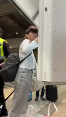A woman with curly hair and bangs smiles and laughs at something behind the camera. She is standing at a train station in front of a train car. She is wearing a white long-sleeved shirt, light gray jeans, and has a black bag slung over one shoulder and a larger off-white bag on the other. She is holding her phone in her left hand. There are other people and luggage behind her.