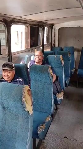 Two men are sitting in an old bus. The seats are blue and have visible wear and tear. The man on the left is wearing a cap and looks directly at the camera and smiles. The man on the right is wearing glasses and also smiles. The bus interior is dated, with visible signs of age. Light enters through the windows. The bus floor is covered with a gray, textured material.