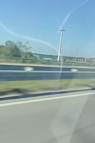 A video captures a view from a moving vehicle. The camera is angled slightly downward, showing a highway with a concrete barrier and a strip of grass. Beyond the barrier, trees and a few buildings are visible. The sky is clear and blue. The movement of the vehicle suggests the camera is recording from inside a car, bus, or train traveling along the highway. The overall scene is a typical roadside landscape under a bright sky.