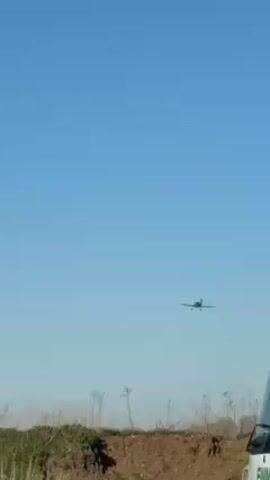 The video shows a small single-engine airplane approaching for landing against a clear blue sky. The plane is dark in color and appears to be flying straight toward the camera. Below, a grassy field with a small earthen bank is visible.