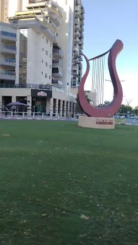 The video shows a statue of a harp in a grassy roundabout. A traffic sign indicating a roundabout is visible in the foreground. Buildings and trees are in the background. The weather appears to be clear and sunny.