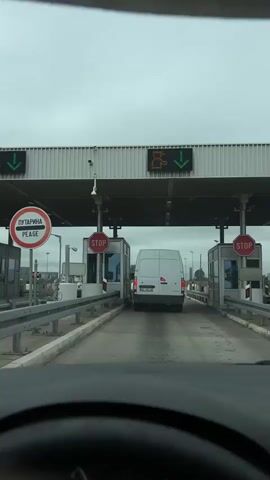 The video shows a view from inside a vehicle approaching a border crossing. Two toll booths are visible, each with a red octagon "STOP" sign. A white van is stopped at one of the booths, and another vehicle is visible further ahead. An overhead display shows an orange icon that appears to be a passport or document being stamped, and a green arrow pointing downwards, indicating the lane is open. The sky is overcast.