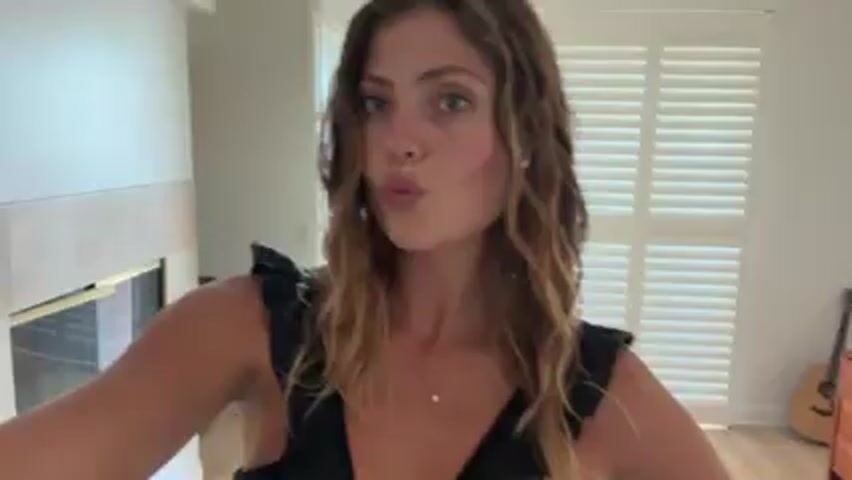 A woman with shoulder-length, wavy, brown hair, wearing a black, low-cut top with ruffles, is facing the camera and making a kissing face. She is indoors, possibly in a home. Behind her, there is a partially visible wall to her left, and a white shuttered window or door to her right, letting in natural light. A small gold necklace is visible on her neck. The scene appears to be casual.