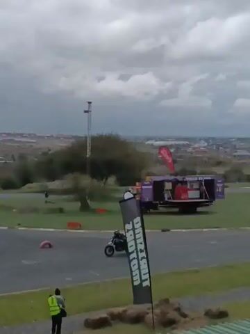 The video shows a go-kart track on a cloudy day. Several motorcyclists ride around the track, some wearing full racing gear. One person in a yellow vest stands near the edge of the track, seemingly monitoring the riders. There is a camera crane next to a white car, suggesting that the event is being filmed. In the distance, a cityscape is visible under the overcast sky. Various flags and cones are placed around the track, marking the turns and boundaries.