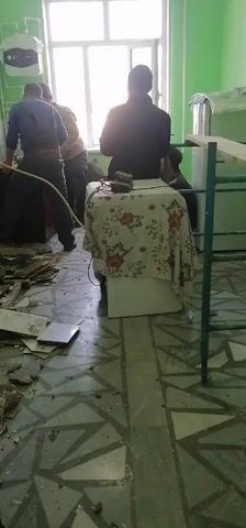 The video shows a group of men working in a room that appears to be undergoing renovation or demolition. The room has light green walls, a patterned tile floor covered with debris, and a large window letting in bright light. One man in a blue shirt is bent over, seemingly working on the floor. Another group of men is clustered around a piece of furniture, possibly a cabinet, which they appear to be moving or working on. The wall behind them shows exposed brick and plaster, indicating that the wall is being taken apart. A table covered with a floral tablecloth sits in the center of the room with a device on top of it. A metal frame, possibly part of a bed, is leaning against the wall.