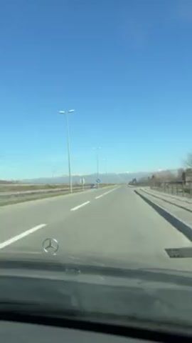 The video shows a car driving on a straight, two-lane road under a clear, blue sky. There are lampposts lining the road and mountains visible in the distance. The car has a Mercedes-Benz emblem on the hood. There is light traffic on the road.