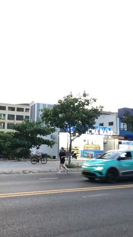 The video captures a street scene in Da Nang, Vietnam. In the foreground, a busy street with yellow double lines indicates traffic. A teal and silver car, possibly an electric vehicle given its design, is driving past, its occupants blurred due to motion. To the right of the car, a motorcycle with a rider in a green shirt is also in motion.

The background features a row of buildings. The most prominent is a store with a bright blue illuminated sign reading "TIỆN LỢI NINA MART" in Vietnamese, meaning "Convenient Nina Mart". The store appears to be a small convenience store or market, with shelves visible through the windows displaying various products. String lights are hung across the storefront, adding to the ambiance, suggesting it's likely late afternoon or early evening. There are tables and chairs set up outside the mart, possibly for customers.

Further to the right, another sign for "NINA MART" is visible, this one with a more traditional sign structure, displaying "CỬA HÀNG TIỆN LỢI NINA MART" (Convenient Store Nina Mart) in blue lettering. The buildings behind the mart are a mix of white and blue, with some trees interspersed.

On the sidewalk in front of the Nina Mart, a person is riding a bicycle. They are wearing a light-colored shirt and a hat. Another motorcycle is visible parked to the left of the store. The overall impression is a lively street scene in an urban environment, likely during daylight or dusk, with commercial activity in full swing. The sky is overcast, suggesting a possibility of rain or simply a cloudy day. The presence of multiple vehicles and pedestrians points to a functioning commercial area.
