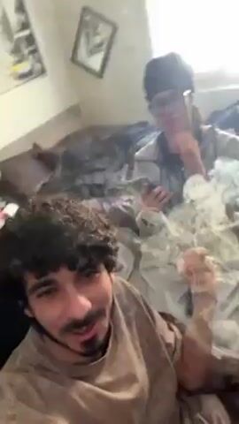 A man with curly hair and a goatee is recording a video in what appears to be a messy living room. He is seated, and another man is standing behind him with his hands clasped in front of his face. There are items scattered on the floor around them. The man in the foreground smiles at the camera and seems to be gesturing to the music playing in the background.