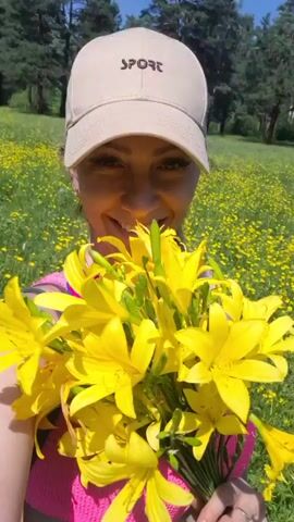 The video features a smiling young woman holding a bouquet of bright yellow flowers. She is wearing a light-colored baseball cap with the word "SPORT" embroidered on the front and a pink top. The background is a field of abundant yellow wildflowers under a clear, sunny sky, suggesting a warm, pleasant day, likely in the late morning or early afternoon. Behind the field, a line of trees indicates a wooded area. The woman's joyful expression and the vibrant flowers create a cheerful and idyllic scene, possibly captured during an outdoor excursion or a visit to a natural setting. The overall impression is one of happiness and appreciation for nature. The location is specified as Dachnaya, Russia.