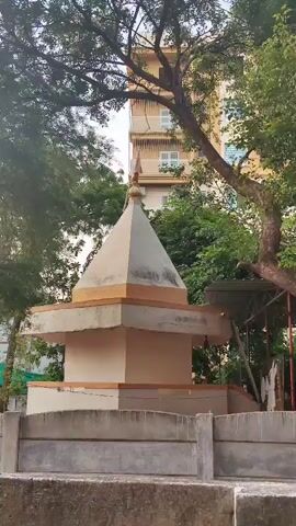 The video shows a close-up view of a small Hindu temple structure. The temple has a tiered base topped with a pyramidal spire. A flag is attached to the very top of the spire. The temple is surrounded by greenery, with trees in the foreground and a tall building visible in the background. The overall scene appears to be outdoors, possibly in an urban or suburban setting.