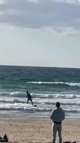 The scene depicts a beach on a partly cloudy day with ocean waves visible. A person is kitesurfing, skillfully maneuvering the kite and board over the waves. The camera focuses on the kitesurfer as they perform aerial tricks, catching air and momentarily rising above the water's surface. In the foreground, another individual stands on the beach, watching the kitesurfer's performance.